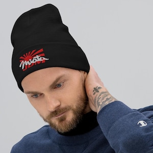 Puede incluir: Gorro negro con la palabra "Miata" bordada y un diseño de sol naciente en rojo y blanco. Este gorro de punto es un accesorio cómodo y moderno, perfecto para cualquier ocasión.