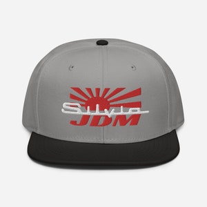Puede incluir: Una gorra de béisbol gris y negra con un sol naciente japonés bordado en rojo y blanco y el texto "SILVIA JDM".