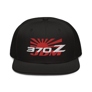 Puede incluir: Gorra de béisbol negra de ala plana con diseño bordado. El diseño presenta un sol naciente rojo con texto blanco y rojo "370Z JDM". La gorra tiene un ajuste estructurado.