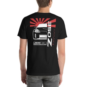 Puede incluir: Camiseta negra con un gráfico blanco de un coche Nissan 350Z, un sol naciente y el texto "Z350" y "JDM 8/17 MANUFACTURED IN JAPAN" en blanco. El texto japonés dice "ニッサン 350Z".