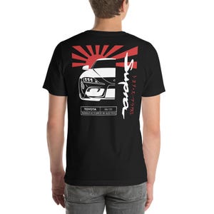 May include: Black t-shirt with a white silhouette of a Toyota Supra car against a red and white Japanese rising sun design. The text "Supra" is written vertically in red and white Japanese characters and the text "TOYOTA  06/70 MANUFACTURED IN AUSTRIA" is written in white below the car.