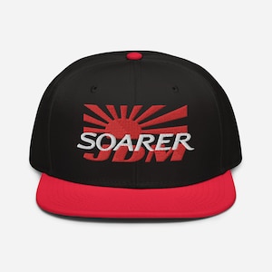 Op de afbeelding: Zwarte en rode baseballpet met een witte geborduurde Japanse opkomende zon en de tekst "SOARER JDM".