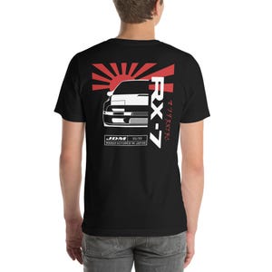May include: Black t-shirt with a white silhouette of a car, a red rising sun, and the text "RX-7" in red and Japanese characters. The text "JDM 85/80 MANUFACTURED IN JAPAN" is also printed on the shirt.