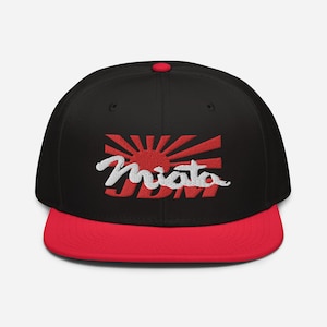 Puede incluir: Gorra de béisbol negra y roja con un logotipo bordado en blanco que dice "Miata JDM" y un gráfico de sol naciente rojo.