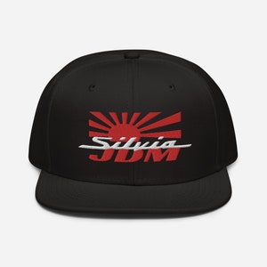 Puede incluir: Gorra de béisbol negra con un gráfico de sol naciente japonés rojo y blanco y el texto "Silvia JDM" bordado en la parte delantera.