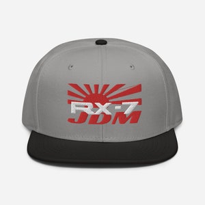 Puede incluir: Una gorra de béisbol gris y negra con un diseño bordado de sol naciente japonés rojo y blanco y el texto "RX-7 JDM".