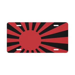 May include: A rectangular license plate featuring a red sun with black rays radiating outwards. The plate has a red background with four mounting holes, and the design is a bold graphic.