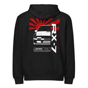 May include: Black hooded sweatshirt featuring a white graphic of a car with the text "RX-7" and Japanese text. The design includes a red sunburst and the text "JDM 05/90 Manufactured in Japan".
