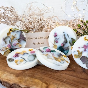 Puede incluir: Cinco fundidos de cera blanca redondos con flores secas y detalles dorados. Los fundidos de cera están dispuestos sobre una superficie de madera. La etiqueta dice "Aroma wax tablet Patchouli".