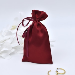 Può includere: Una borsa di raso rosso con coulisse e un nastro bianco. La borsa è appoggiata su una superficie bianca con orecchini a cerchio dorati accanto.