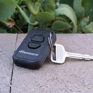 May include: A black key fob with two buttons and the word "Runner" printed on it, attached to a silver key. The key has the Toyota logo. The key and fob are on a concrete surface.