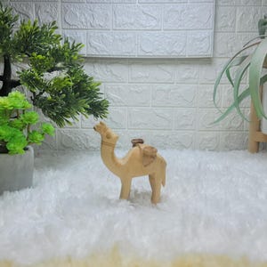 Peut inclure: Petite figurine de chameau en bois clair, avec une selle. Le chameau est debout sur quatre pattes. En arrière-plan, des plantes vertes et un mur de briques blanches.