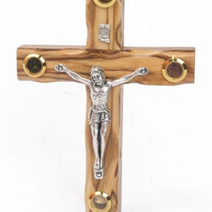 May include: A wooden crucifix with a silver Jesus figure. The cross is made of light brown wood and features four circular insets with gold-colored rims, each containing a different colored stone. The top of the cross has a small silver emblem.