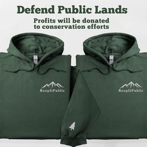 May include: Two dark green hooded sweatshirts with white mountain and text graphics. The text reads "Keep It Public". The text "Defend Public Lands" and "Profits will be donated to conservation efforts" is displayed above the sweatshirts.