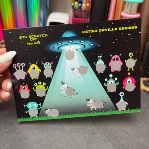 Op de afbeelding: Een zwarte en groene kraskaart met een UFO, koeien en aliens. De kaart vermeldt "€ 7,50 Kras € 9 Maximum" en "Patina DeVille Designs".