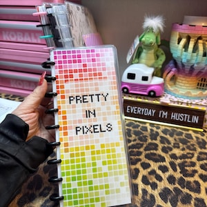 以下が含まれることがあります： カラフルなピクセルデザインと「PRETTY IN PIXELS」というテキストが書かれた透明なプラスチックカバー。カバーは、小さな黒いリングバインダーノート用です。
