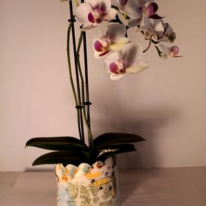 Puede incluir: Una orquídea en maceta con flores blancas y moradas. La planta está en una maceta de cerámica con borde festoneado, decorada con un diseño pintado a mano de cactus y otras plantas en una variedad de colores. La maceta mide aproximadamente 15 cm de alto.