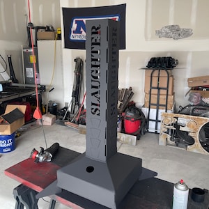 May include: A black metal fire pit with the word "SLAUGHTER" cut out on the side. The fire pit is sitting on a wooden table in a garage.