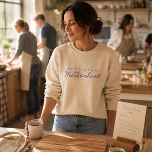 Pode incluir: Uma camisola bege de gola redonda com o texto roxo "executive dysfunction Motherhood" estampado no peito. A peça tem mangas compridas e gola, punhos e bainha canelados, usada num ambiente de cozinha com vários ingredientes culinários.