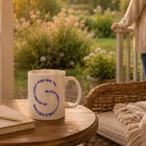 May include: A white ceramic mug with a handle and blue text arranged in a spiral pattern that reads, Please don't ask me to make another decision. The mug sits on a round wooden table next to a closed notebook with a gold pen resting on top.