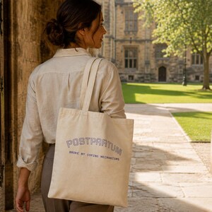 May include: A cream-colored canvas tote bag with two shoulder straps, featuring the dark blue text POSTPARTUM BROKE MY COPING MECHANISMS printed in a bold, arched font on the front surface of the bag, which is carried over the shoulder.