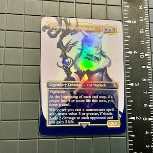 May include: A holographic trading card featuring a cat-like character named Y'shtola, Night's Blessed. The card has a blue border and includes text describing the character's abilities. A ruler is next to the card.