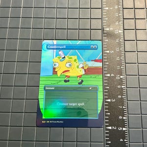 May include: A holographic trading card featuring SpongeBob SquarePants as the central character. The card is labeled "Counterspell" with the text "Counter target spell." The card has a blue and green color scheme.