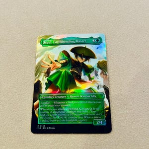 May include: A holographic trading card featuring "Toph, Earthbending Master." The card depicts a character in green attire with a green hat, set against a blurred background. The card includes text describing the character's abilities and stats, with a 2/4 power and toughness.