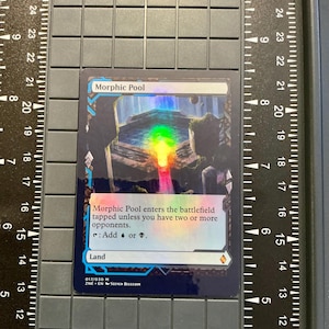 May include: A holographic "Morphic Pool" trading card. The card features a landscape with a rainbow-colored light beam. Text on the card reads "Morphic Pool" and game instructions. The card is a collectible item.
