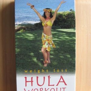 Peut inclure: Une jaquette de DVD pour une vid&eacute;o d'entra&icirc;nement de hula avec une femme en jupe florale jaune et orange et un haut de bikini jaune. La jaquette est blanche avec du texte rouge qui dit "HULA WORKOUT with Kili" et "weight loss". L'arri&egrave;re-plan est une zone herbeuse verte avec une mer bleue au loin.