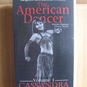 Peut inclure: Une cassette vid&eacute;o VHS avec un fond noir et un titre rouge qui dit "The American Dancer: Volume 1 - Cassandra Introduction to Bellydancing". La couverture pr&eacute;sente une femme en costume noir et blanc ex&eacute;cutant une danse du ventre.