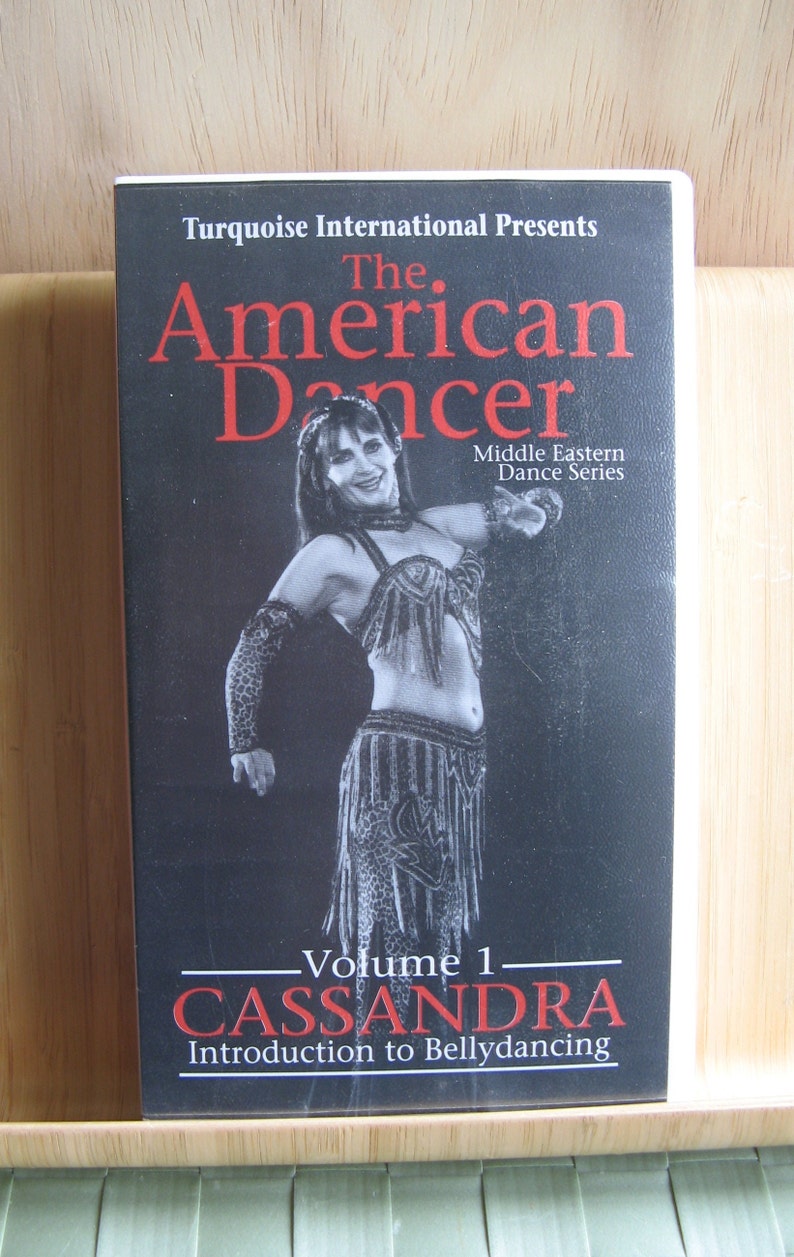 Peut inclure: Une cassette vid&eacute;o VHS avec un fond noir et un titre rouge qui dit "The American Dancer: Volume 1 - Cassandra Introduction to Bellydancing". La couverture pr&eacute;sente une femme en costume noir et blanc ex&eacute;cutant une danse du ventre.