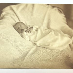 May include: A black and white photograph of a baby sleeping in a white blanket in a wicker chair.