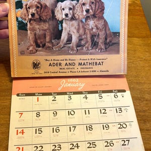 May include: Vintage 1962 calendar featuring a color illustration of three Cocker Spaniel puppies. The calendar includes the month of January and real estate and insurance advertising.