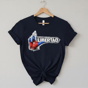 Puede incluir: Camiseta azul marino con un diseño de gesto de la mano con los colores de la bandera cubana y la palabra "LIBERTAD". El diseño tiene un aspecto desgastado y vintage. La camiseta está colgada en una percha de madera.