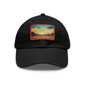 May include: Black baseball cap with a rectangular patch featuring a desert landscape painting. The patch has a brown border and depicts a canyon scene with blue, yellow, and brown hues. The cap is made of a soft material.