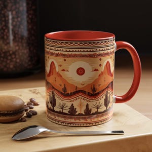 以下が含まれることがあります： 砂漠の風景のデザインが施された赤いセラミック製のマグカップ。デザインには、山、サボテン、幾何学模様の太陽が描かれています。マグカップには赤い取っ手があります。