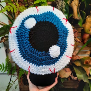 May include: A crocheted eye with a blue iris and white sclera. The eye is surrounded by a white crocheted border with red stitching.