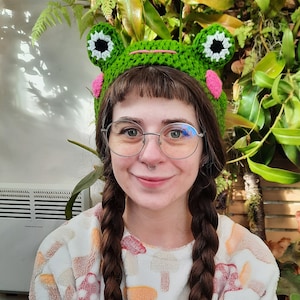 Op de afbeelding: Een vrouw met lang gevlochten haar draagt een groene, gebreide muts in de vorm van een kikker met witte en roze accenten. Ze draagt een wit shirt met een paddenstoelprint.
