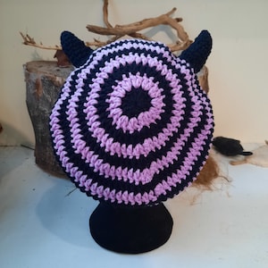 Puede incluir: Un gorro de punto morado y negro con cuernos. El gorro tiene un diseño circular con rayas alternas de hilo morado y negro.