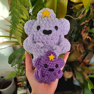 May include: Two plush cloud-shaped figures made of yarn. The larger figure is light purple with a yellow felt star on its head, while the smaller figure is a darker purple with a matching yellow star. Both have black plastic eyes and mouths.