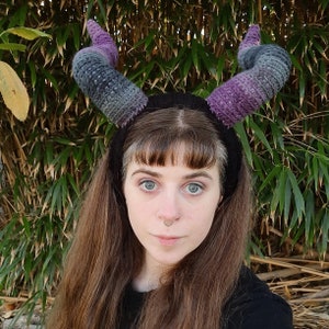 Puede incluir: Una persona lleva una diadema negra con cuernos de crochet morado y gris. La diadema se lleva sobre el pelo y los cuernos apuntan hacia arriba.