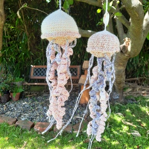 May include: Two crocheted jellyfish ornaments hanging from a tree branch. The jellyfish are made with a variety of colors, including pink, white, and purple. The jellyfish are hanging from a tree branch with green leaves.