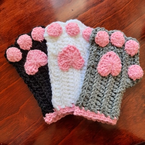 May include: Three crocheted paw-shaped fingerless gloves in black, white, and gray with pink paw pads.