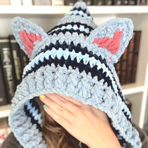 May include: A handmade, light gray crochet hood with cat ears. The hood features stripes of navy blue and light blue. The inner ears are a coral pink color. The hood is designed to be worn, with a pointed top and a face opening.