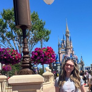 Może przedstawiać: Kobieta w szarej koszulce i okularach z niebiesko-r&oacute;żową opaską Minnie Mouse stoi przed Zamkiem Kopciuszka w Walt Disney World. Zamek jest ozdobiony niebieskimi i r&oacute;żowymi wieżami i ma biały dach. Na pierwszym planie wiszą r&oacute;żowe kwiaty z latarni.