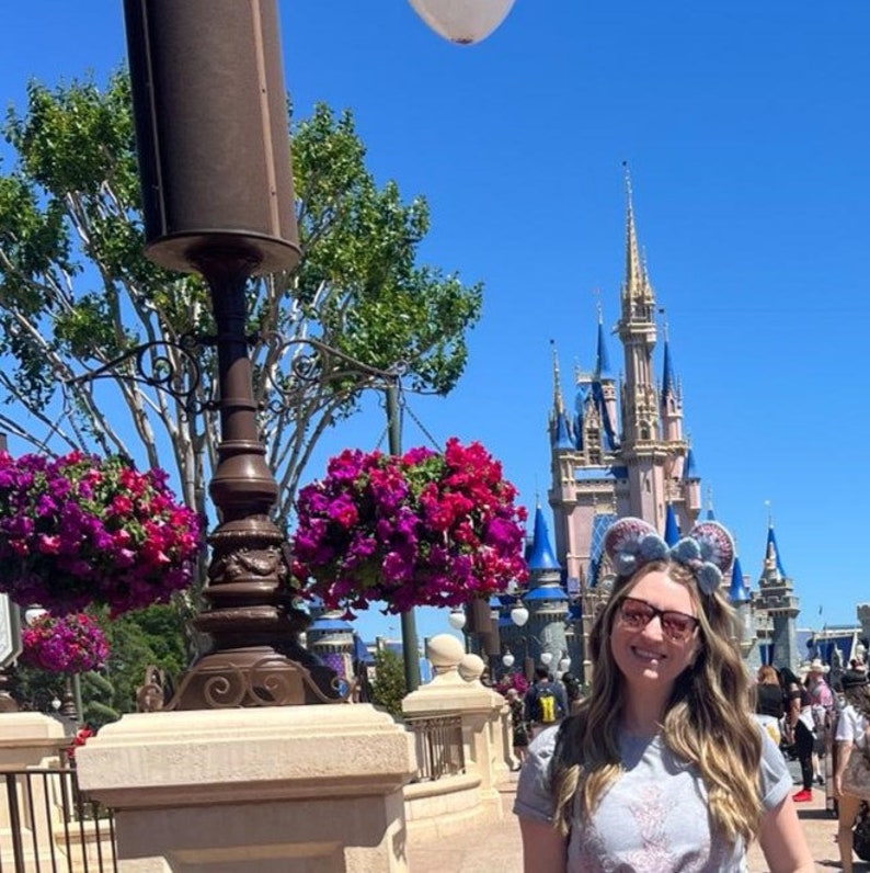 Może przedstawiać: Kobieta w szarej koszulce i okularach z niebiesko-r&oacute;żową opaską Minnie Mouse stoi przed Zamkiem Kopciuszka w Walt Disney World. Zamek jest ozdobiony niebieskimi i r&oacute;żowymi wieżami i ma biały dach. Na pierwszym planie wiszą r&oacute;żowe kwiaty z latarni.