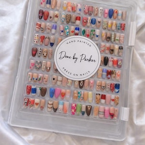 May include: A clear plastic container filled with an assortment of hand-painted press-on nails. The nails feature various designs, including hearts, stripes, and floral patterns. The container has a circular label that reads "Done by Parker, Hand Painted, Press on Nails."