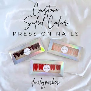 May include: Three boxes of custom solid color press-on nails are displayed. One box contains dark burgundy nails, another has a nude pink shade, and the third features bright red nails. Each box has a holographic border and a circular sticker with the brand name.