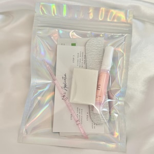 May include: A clear, iridescent zip-lock bag containing nail application supplies. The kit includes a nail file, alcohol prep pad, cuticle stick, nail glue, and instructions. The text "Nail Application" is visible on the instructions.
